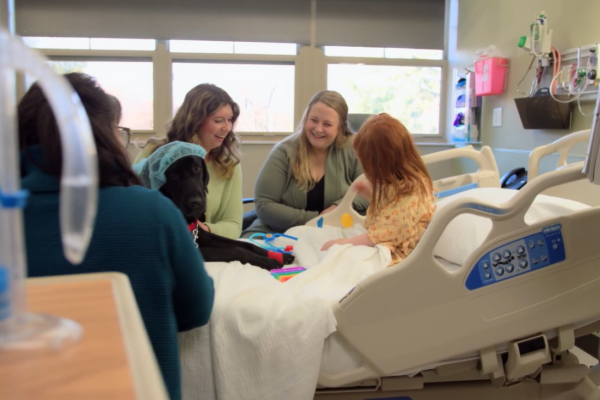 three adults smiling while child plays with toys in hospital bed