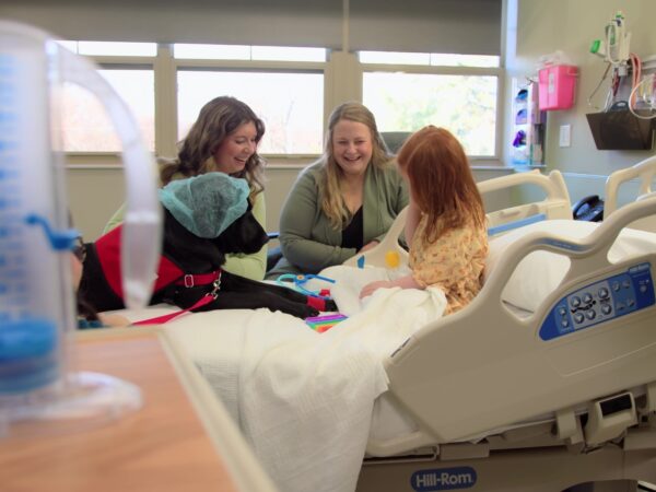 three adults smiling while child plays with toys and dog in hospital bed