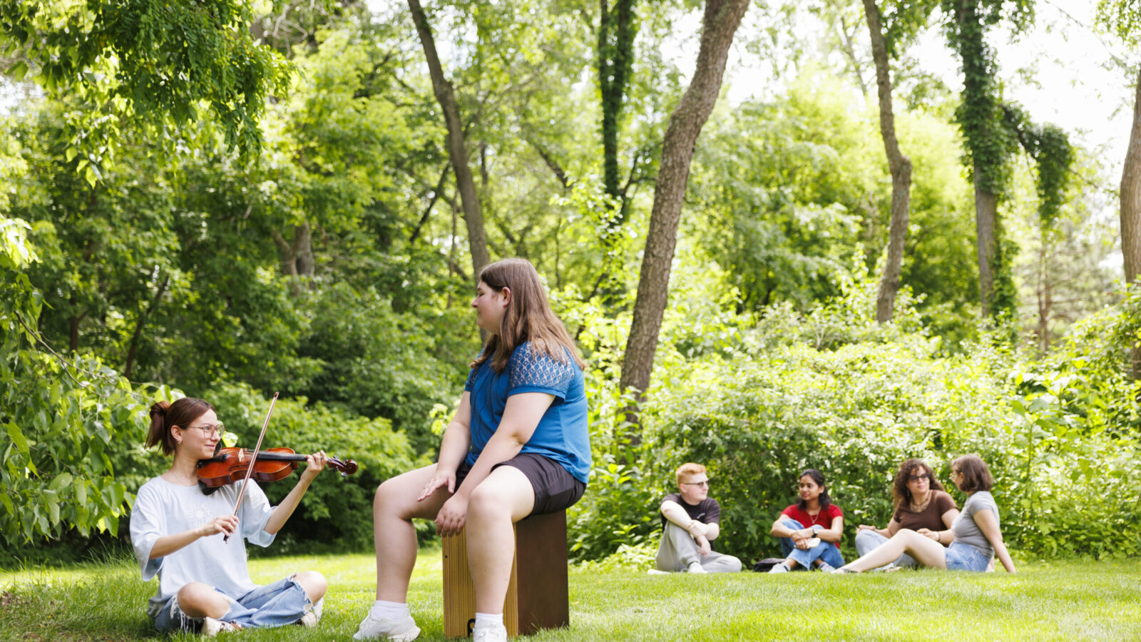 Woman playing violin while students in the background are sitting in a circle
