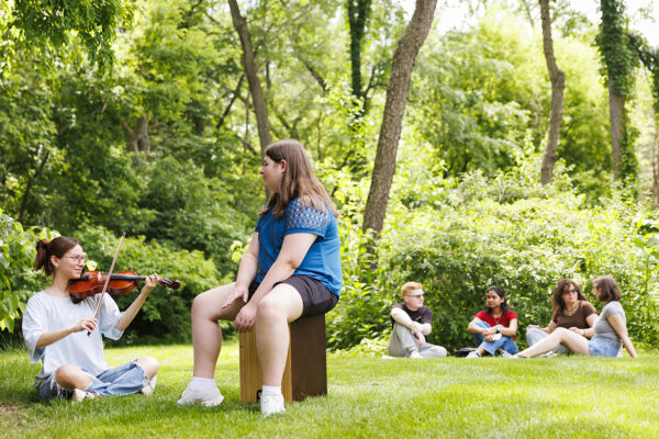 Woman playing violin while students in the background are sitting in a circle